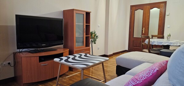 Un salón con televisor de pantalla plana, aparador de madera, tabla de planchar, sofá y una mesa de comedor con sillas.