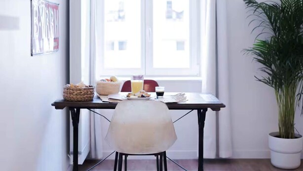 Un comedor con una mesa preparada para dos personas, una planta en maceta y un ventanal con vistas a edificios.