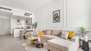 A modern living room with a beige sofa, a coffee table, and a dining area with a white kitchen in the background. - Silkhaus Belgravia Heights, JVC Dubai (Dubai)