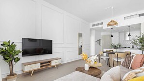 A modern living room with a flat-screen TV, a sofa, a coffee table, and a dining area in the background. - Silkhaus Belgravia Heights, JVC Dubai (Dubai)