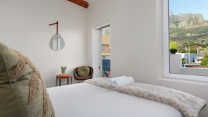 A bedroom with a bed, a small table with a plant, a chair, and a view of a mountain through a window.