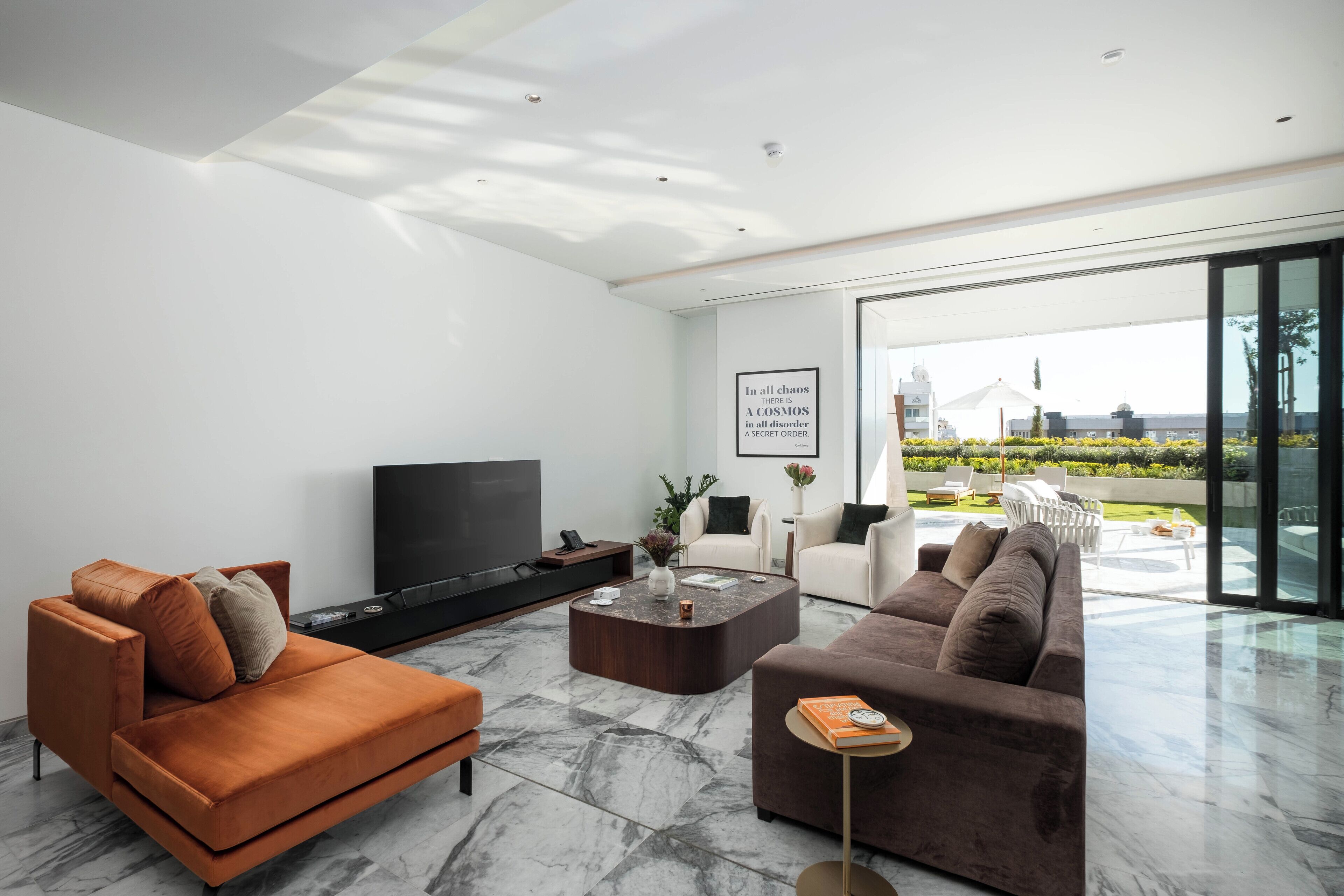 A modern living room with a large flat-screen TV, two sofas, a coffee table, and a large window offering a view of a garden and a building.