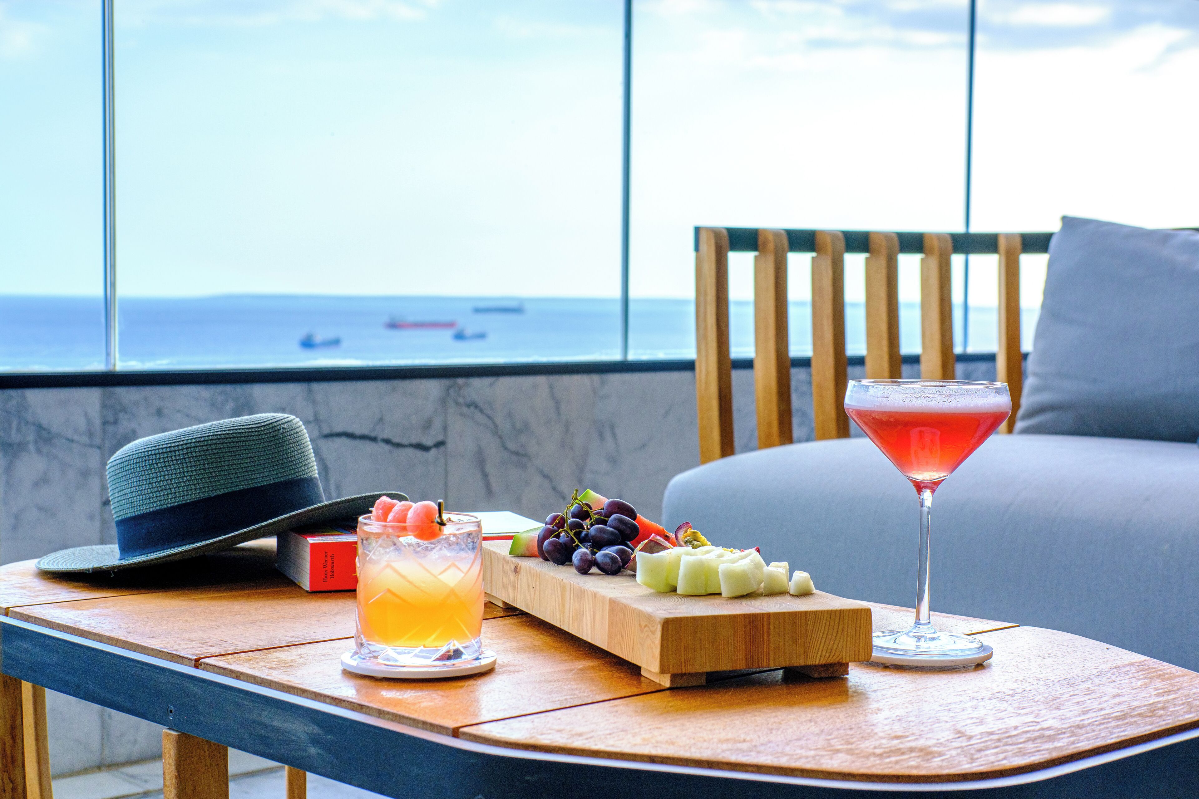 A table with a hat, a tray of fruits, a cocktail, and a glass of orange juice on a balcony with a view of the sea.