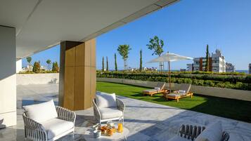 Un patio moderno al aire libre con mobiliario de mimbre blanco, suelo de mármol y vista al mar y edificios al fondo.
