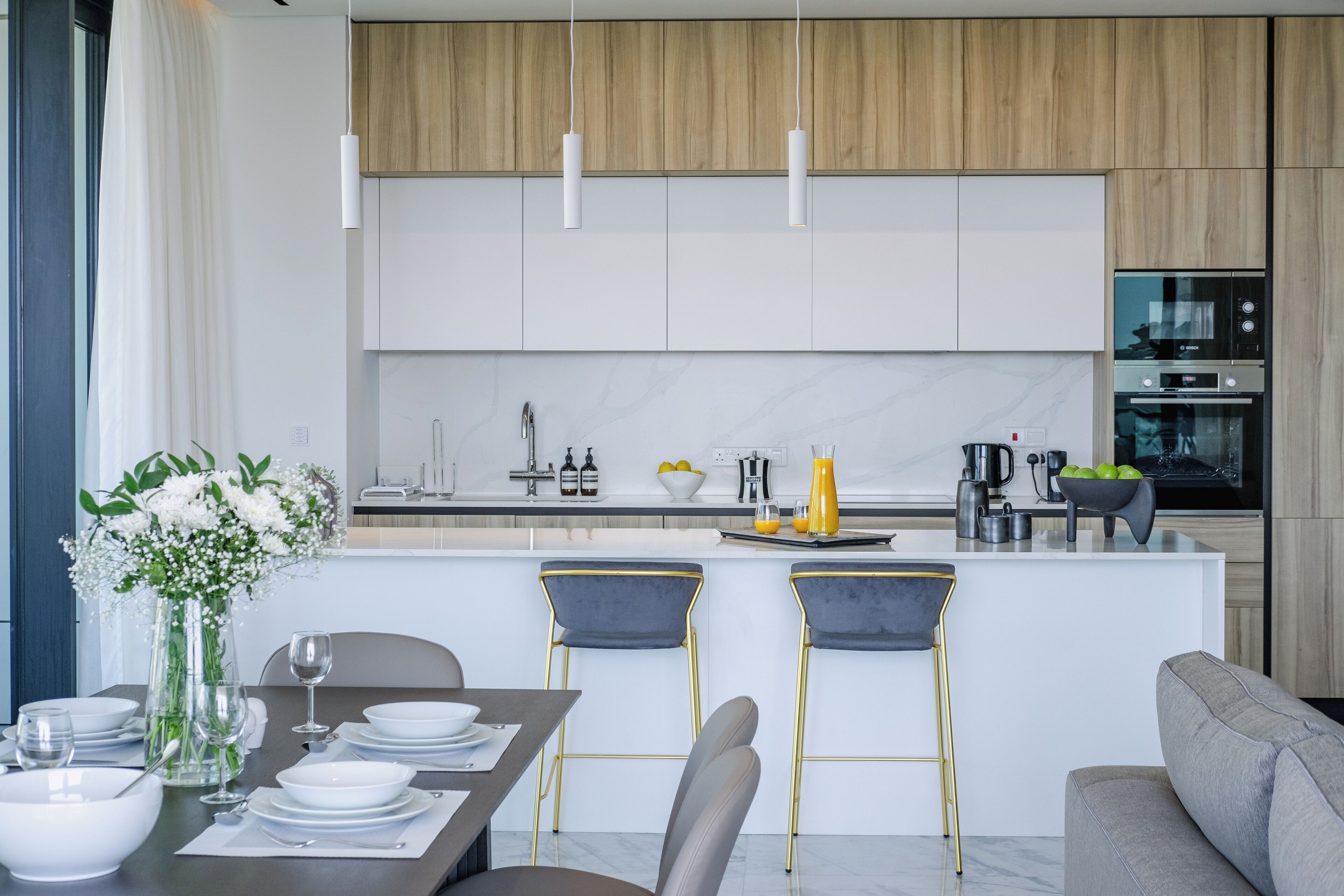 Uma cozinha moderna com armários brancos, bancada de mármore e uma ilha central com banquetas.
