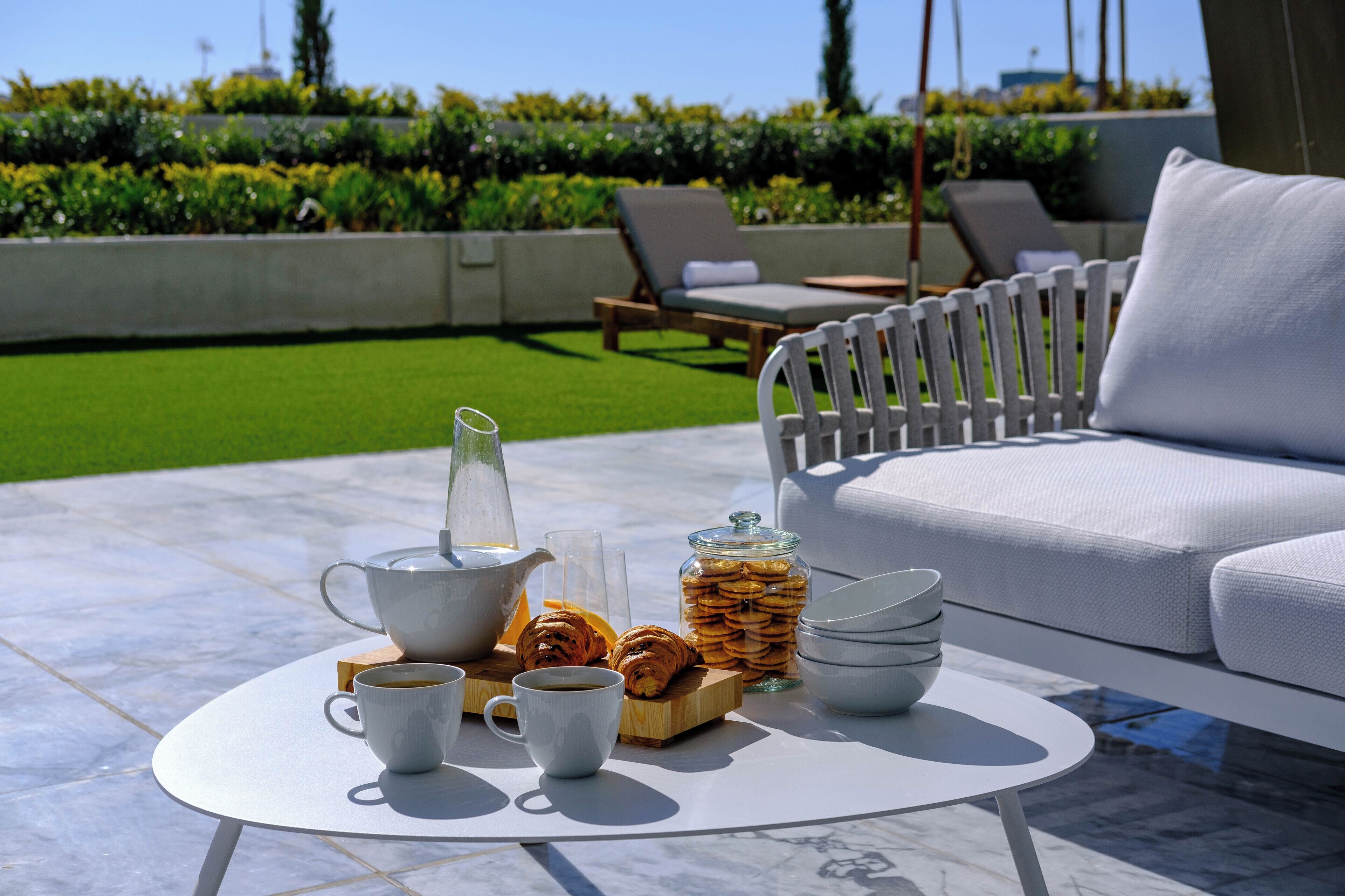 A breakfast setup with a coffee pot, cups, pastries, and a jar of cookies on a round table.