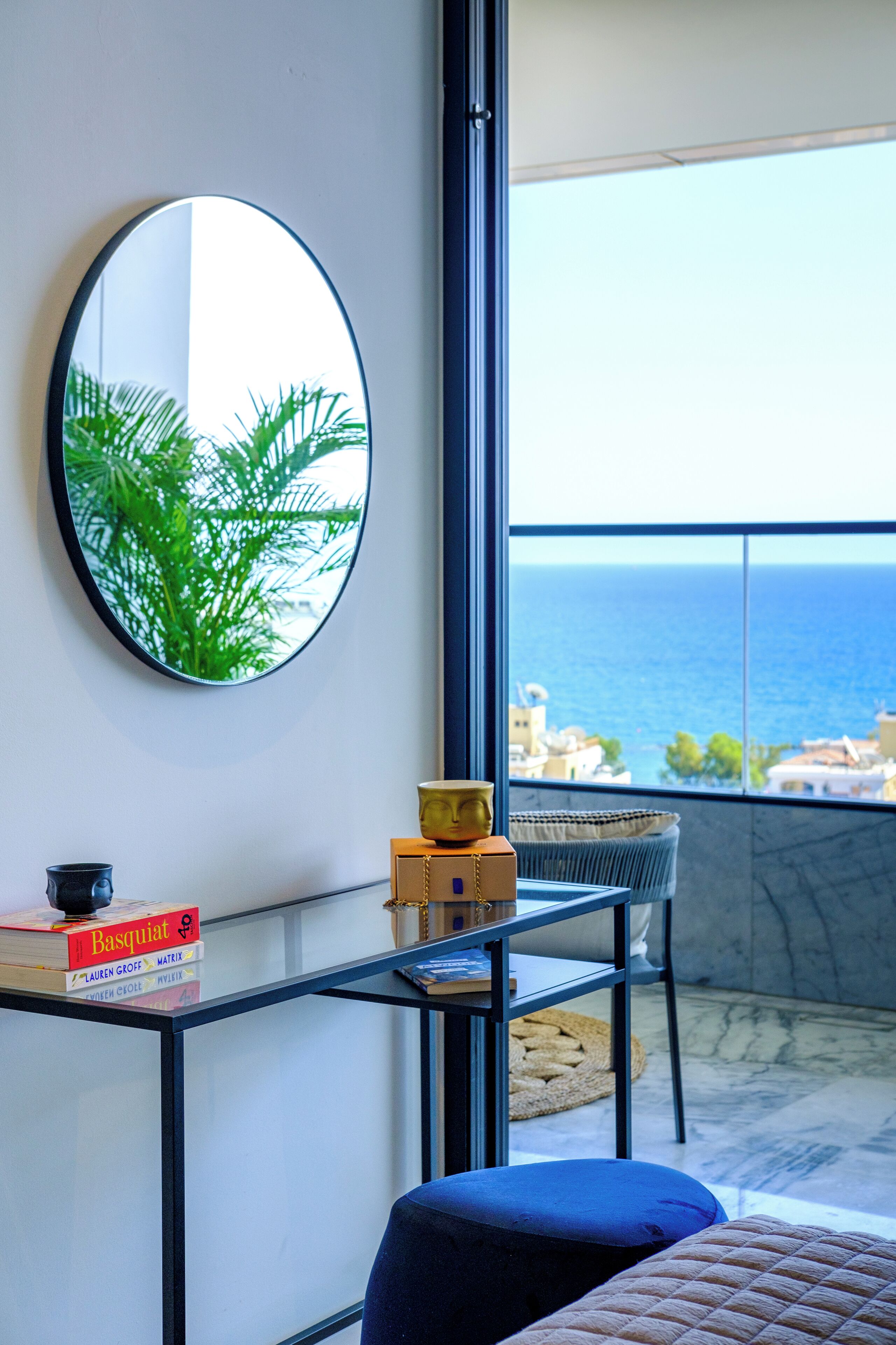 Una habitación con un espejo redondo, una mesa de vidrio con libros, un cojín azul y vistas al mar.