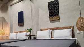 Een tweepersoonsbed met een donkergrijze spreidekker, twee witte kussens en een houten hoofdbord. Er staat een klein tafeltje met een plantje en een wandlamp.