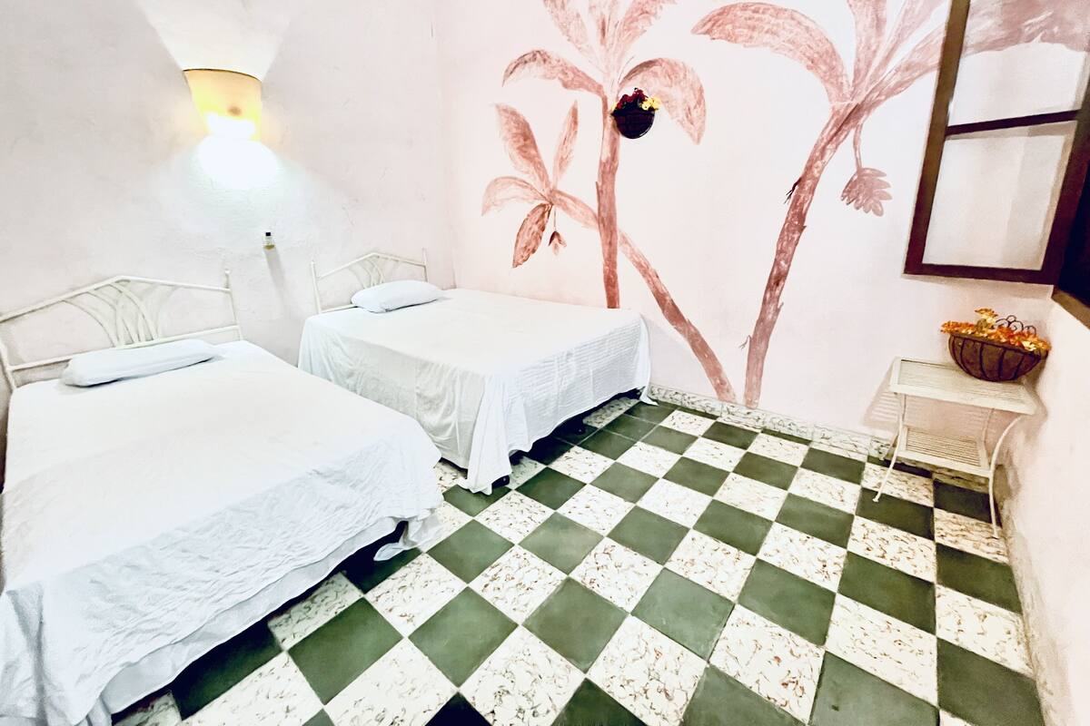 A room with two single beds, a checkered floor, a wall mural, and a small table with a basket.