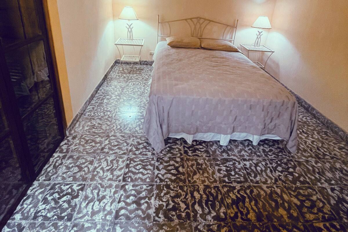 A small, dimly lit room with a patterned floor, a single bed with a headboard, and two bedside lamps.