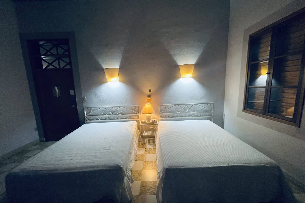 A room with two single beds, a nightstand with a lamp, a door, and a window with wooden shutters.