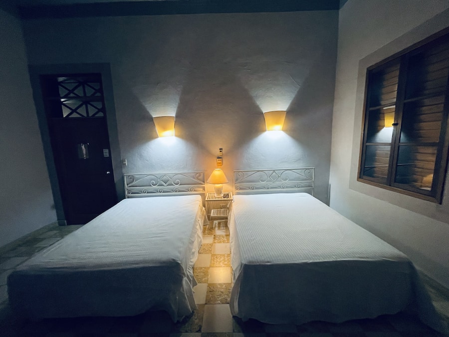 A room with two single beds, a nightstand with a lamp, a door, and a window with wooden shutters.