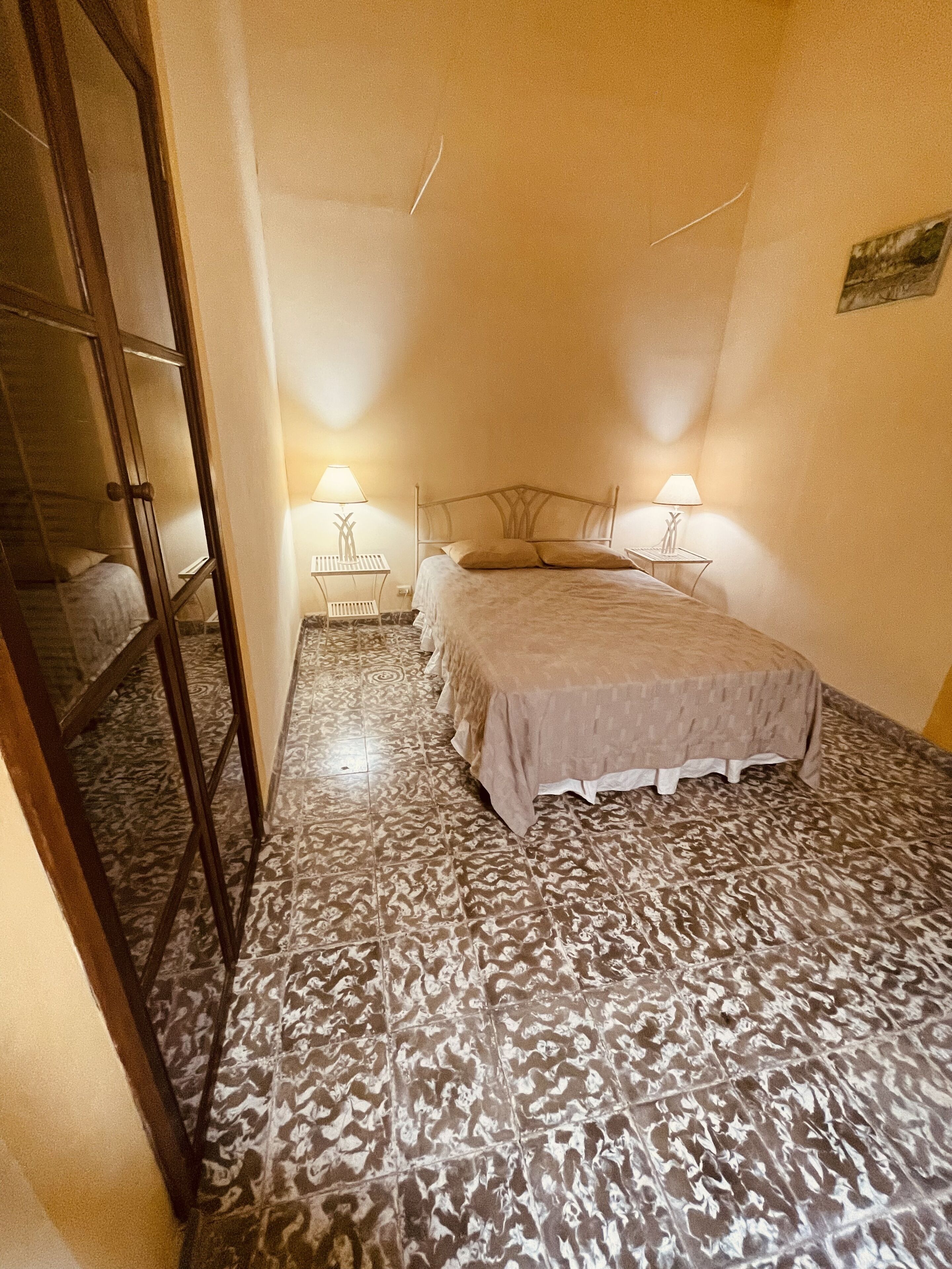 Een slaapkamer met een vloer met patroon, een bed met een witte sprei, twee nachtlampjes en een spiegel aan de muur.