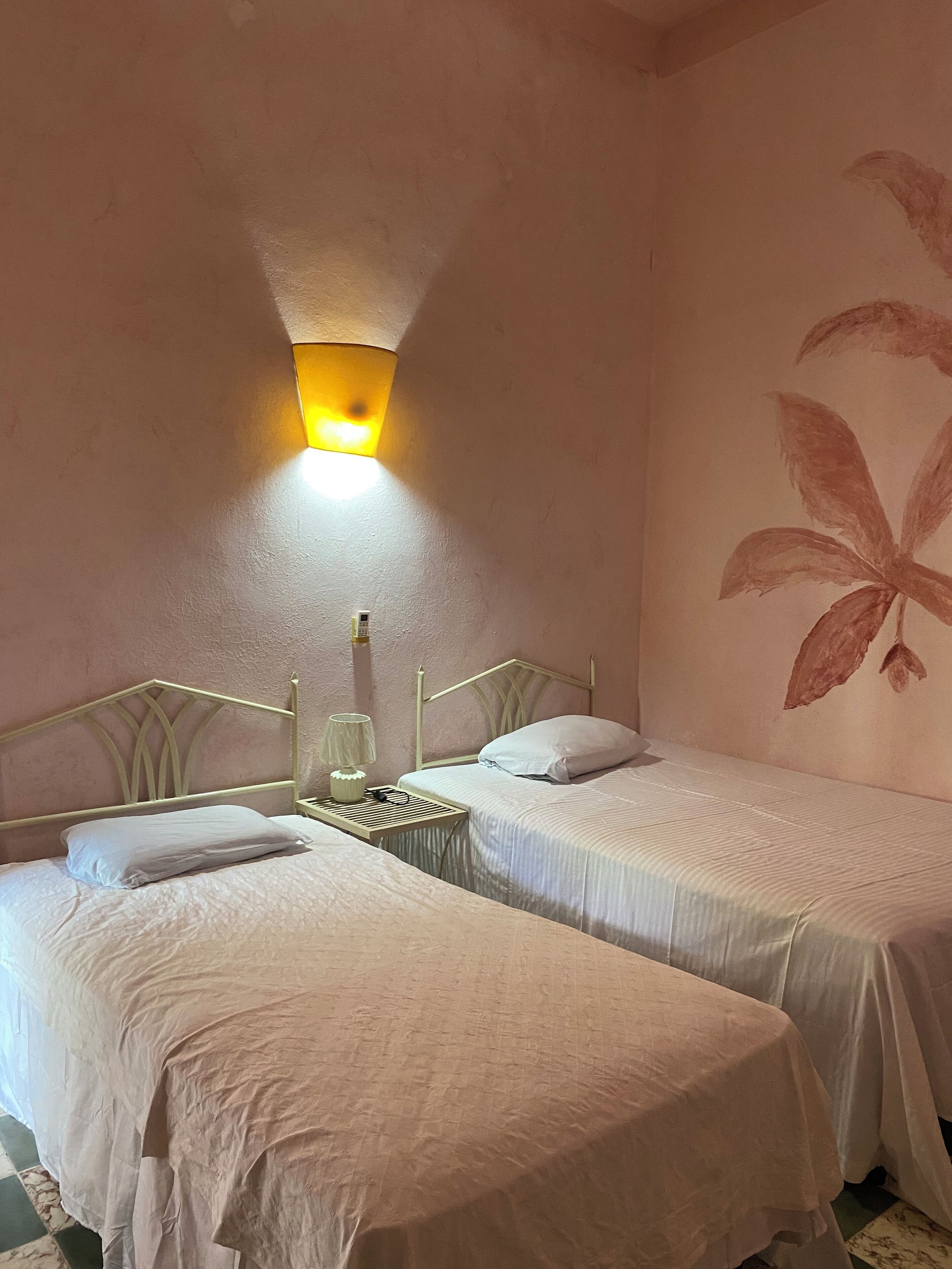 A room with two single beds, a nightstand with a lamp, and a wall-mounted light fixture.