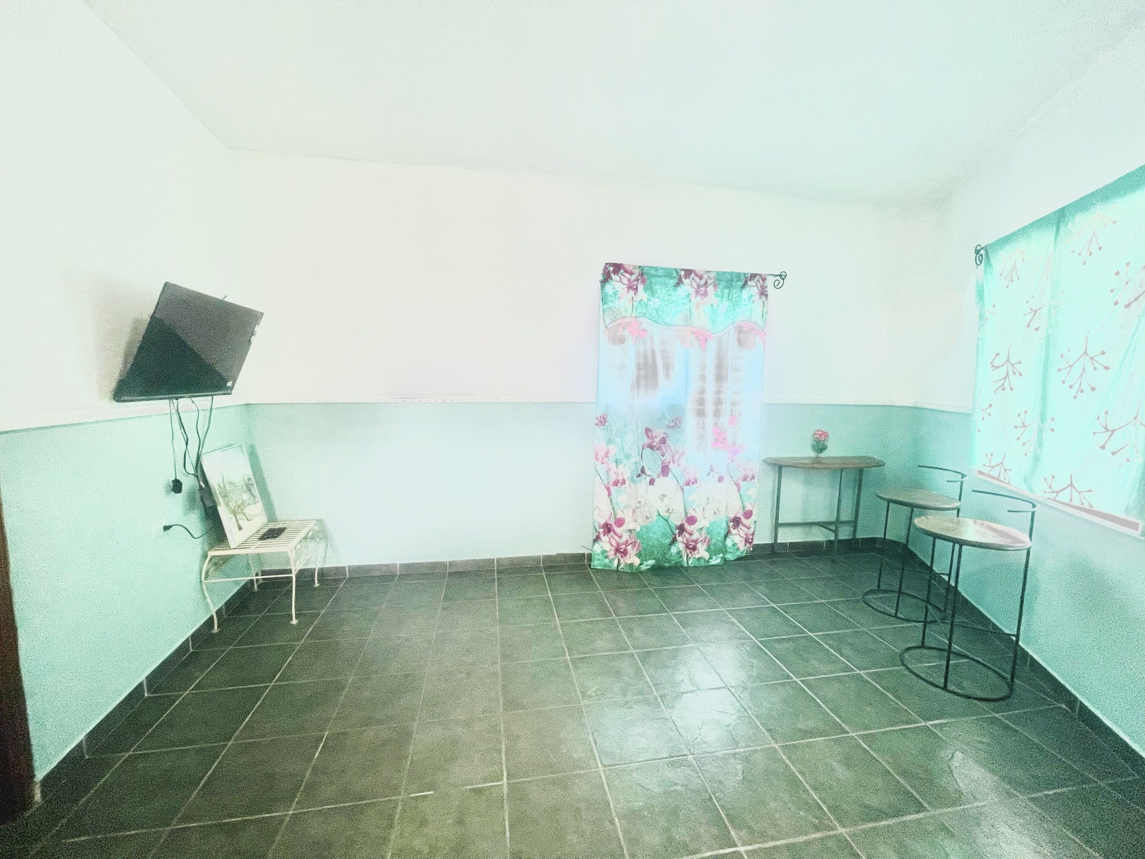 A room with a tiled floor, a chair, a small table, a TV mounted on the wall, and a curtain with a floral pattern.