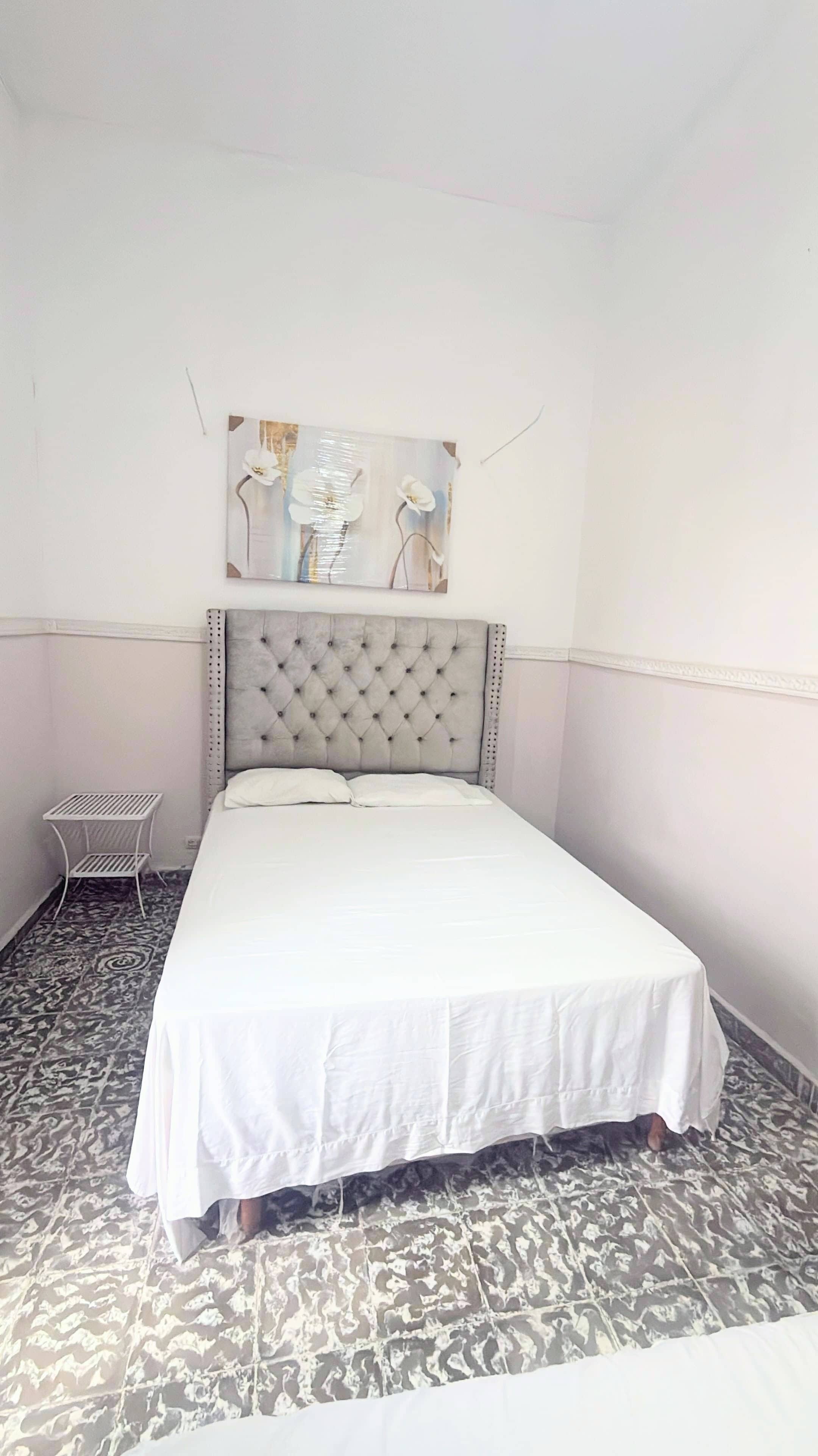 Une chambre à coucher avec un lit, une tête de lit blanche, une petite table de chevet blanche et un tableau accroché au mur.