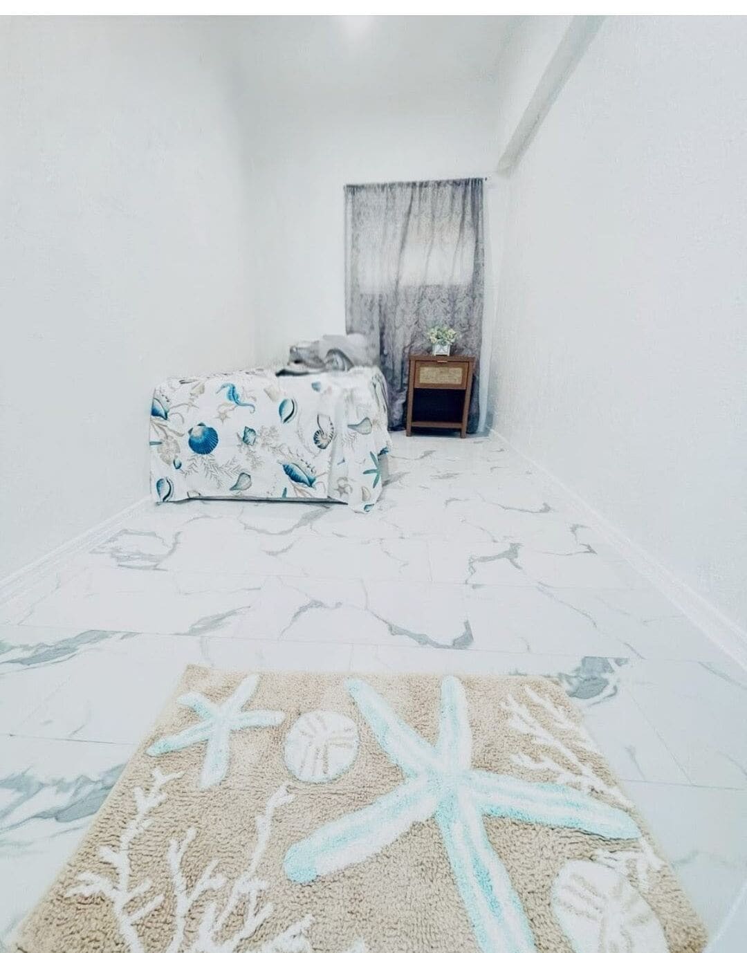 A bedroom with a bed, a nightstand, a window with curtains, and a starfish-patterned rug.