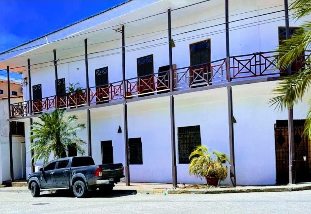 Un edificio de dos pisos con fachada blanca, barandas rojas y un vehículo estacionado.