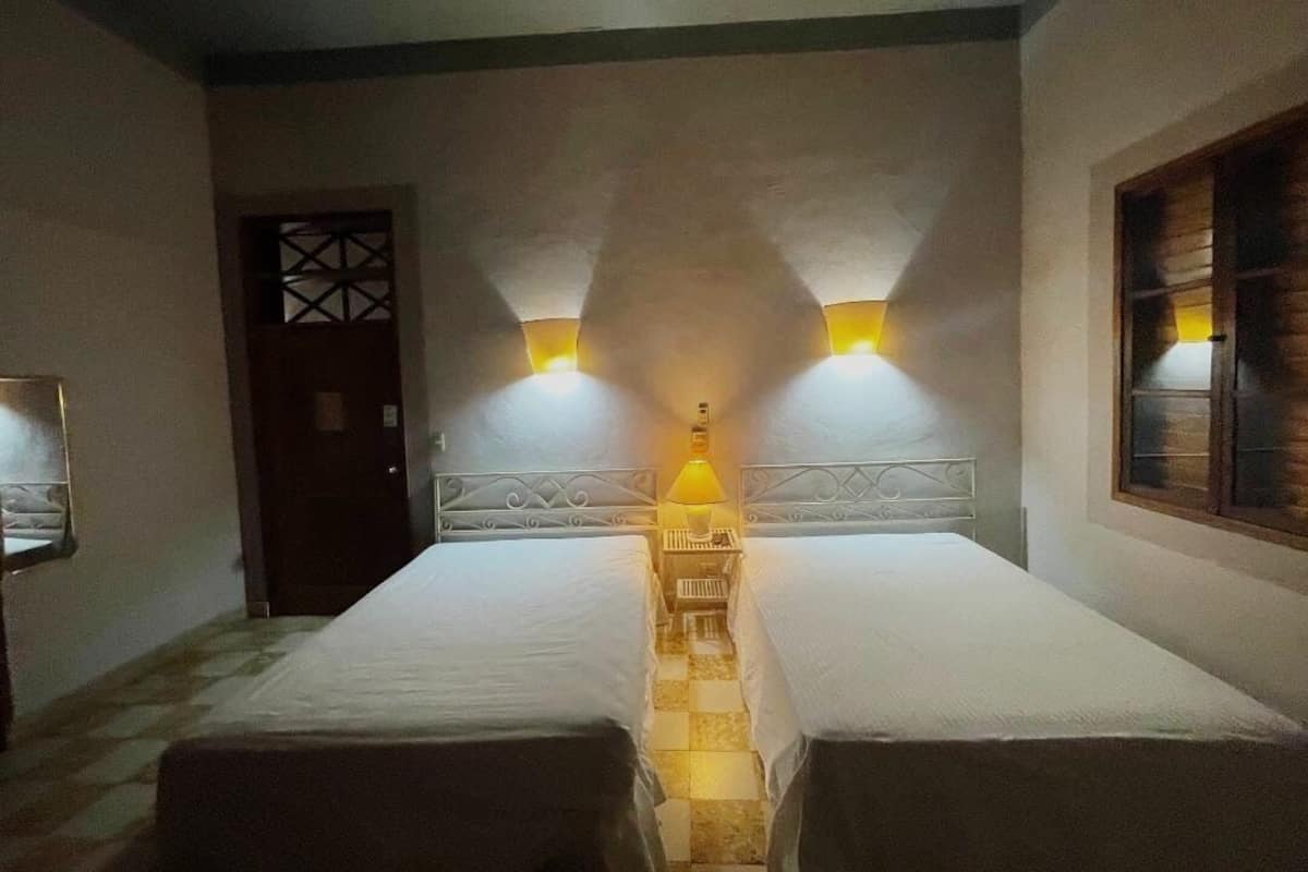 A room with two beds, a nightstand, a wall-mounted lamp, and a window with blinds.