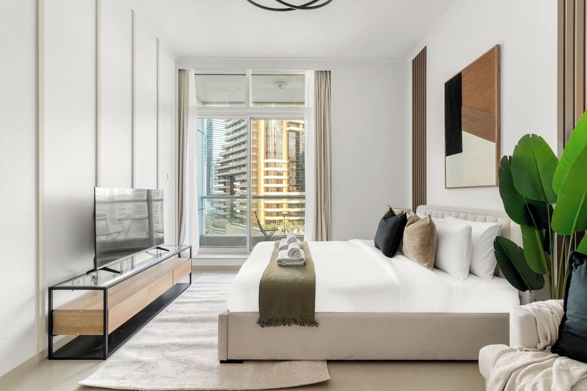 A modern bedroom with a large bed, a TV on a glass cabinet, and a view of tall buildings through the window.