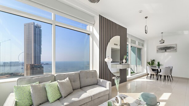 A modern living room with a large window, a sofa, a dining area, and a view of a tall building. - Silkhaus Botanica Tower, Dubai Marina (Dubai)