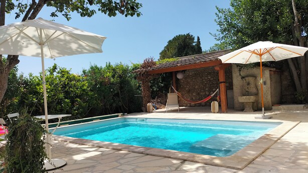 Une piscine agrémentée de deux parasols blancs, entourée de dalles de pierre et de verdure.