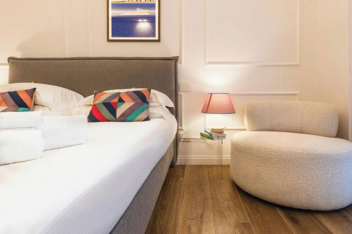 Habitación de hotel con cama, una silla, una mesita de noche con lámpara y un cuadro en la pared.