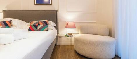 Habitación de hotel con cama, una silla, una mesita de noche con lámpara y un cuadro en la pared.