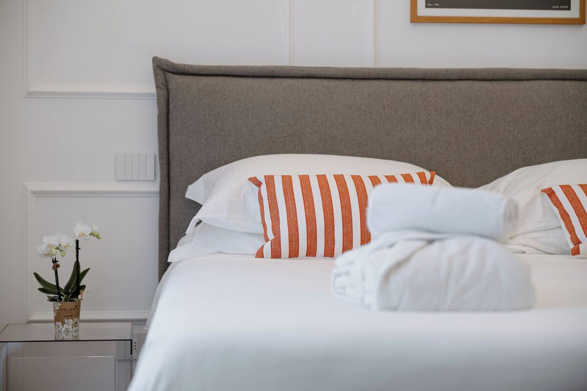 Una cama bien hecha con almohadas a rayas y una toalla doblada, un pequeño jarrón con flores y un cuadro en la pared.