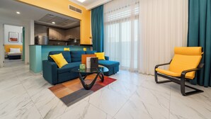 A modern living room with a sofa, armchair, and coffee table, a kitchen area, and a large window with curtains. - izzzi life Mint Holiday Home Apartments (Dubai)