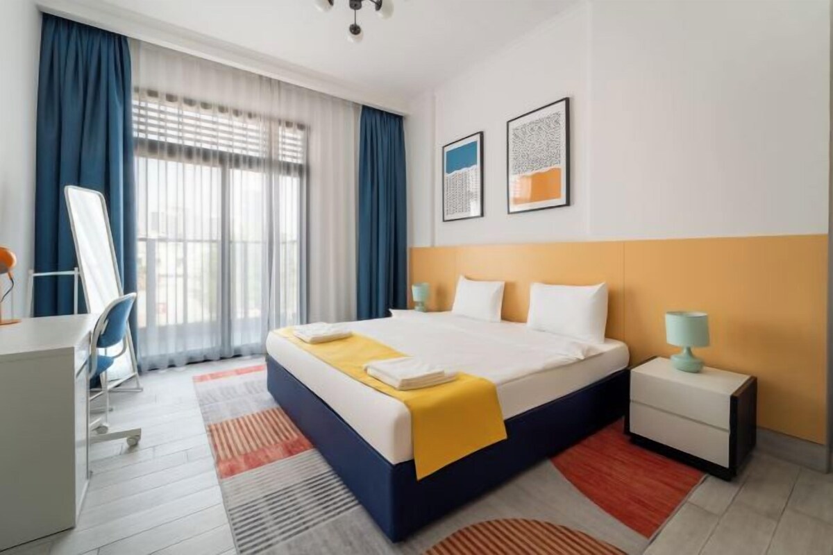 Habitación de hotel con una cama grande, un escritorio, una silla, una mesita de noche y una ventana con cortinas.