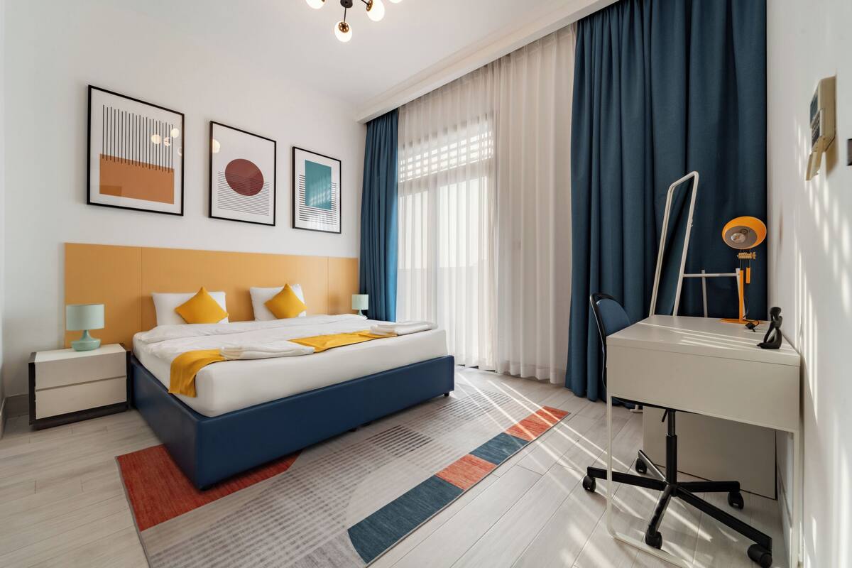 A modern hotel room with a bed, bedside table, lamp, and a desk with a chair.
