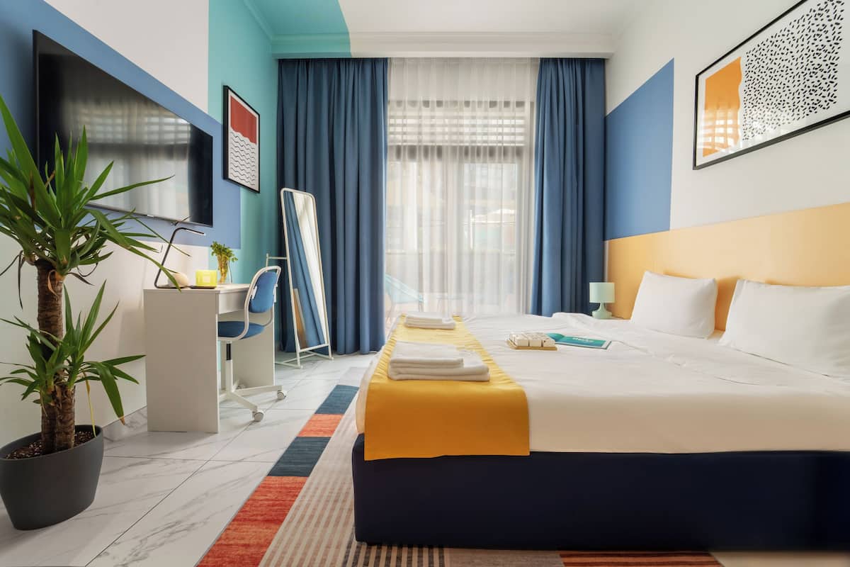 A hotel room with a bed, a desk, a chair, a potted plant, and a window with curtains.
