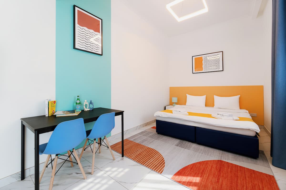 A modern hotel room with a bed, desk, chairs, and artwork on the walls.