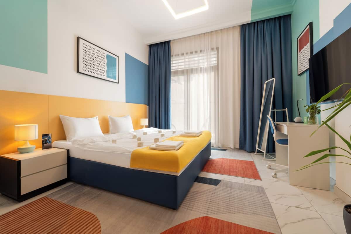 A modern hotel room with a bed, bedside table, desk, chair, and a potted plant.