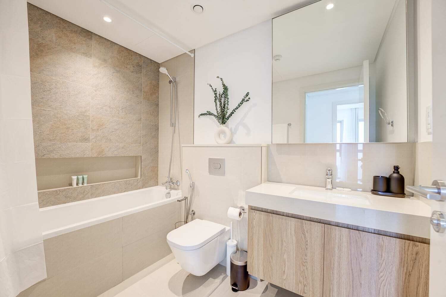 A modern bathroom with a bathtub, a toilet, and a sink. There is a mirror above the sink and a plant on the wall.