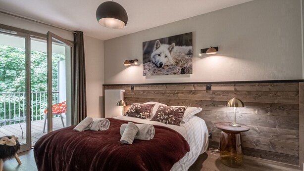 A modern bedroom with a large bed, a wooden headboard, a painting of a dog, and a balcony with a view of trees.