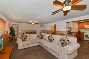 Ein Wohnzimmer mit Ecksofa, Deckenventilator, Fernseher und Küchenzeile mit Holzausstattung und Geräten.