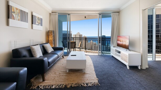 A modern living room with a black leather sofa, a white coffee table, and a TV on a white stand. There are abstract paintings on the wall and a view of the city through large glass doors.