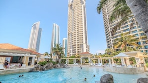 A resort pool area with a clear view of tall buildings in the background, a lounge area with umbrellas, and a rocky pool edge.