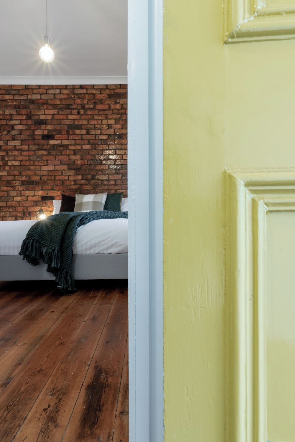 Une chambre avec un mur en briques, un lit avec une literie blanche, un plaid vert et un luminaire suspendu.