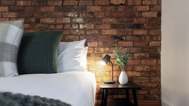 A neatly made bed with white bedding and a green knitted pillow, a small bedside table with a lamp and a vase of flowers, and a brick wall in the background.