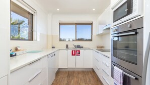 A modern kitchen with white cabinetry, a built-in oven, and a window with a view of palm trees. - Braemar 7 - Lakefront apartment (Forster)