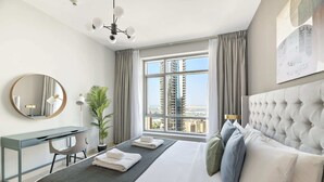 A hotel room with a large bed, a desk, a chair, a potted plant, and a view of tall buildings through the window.