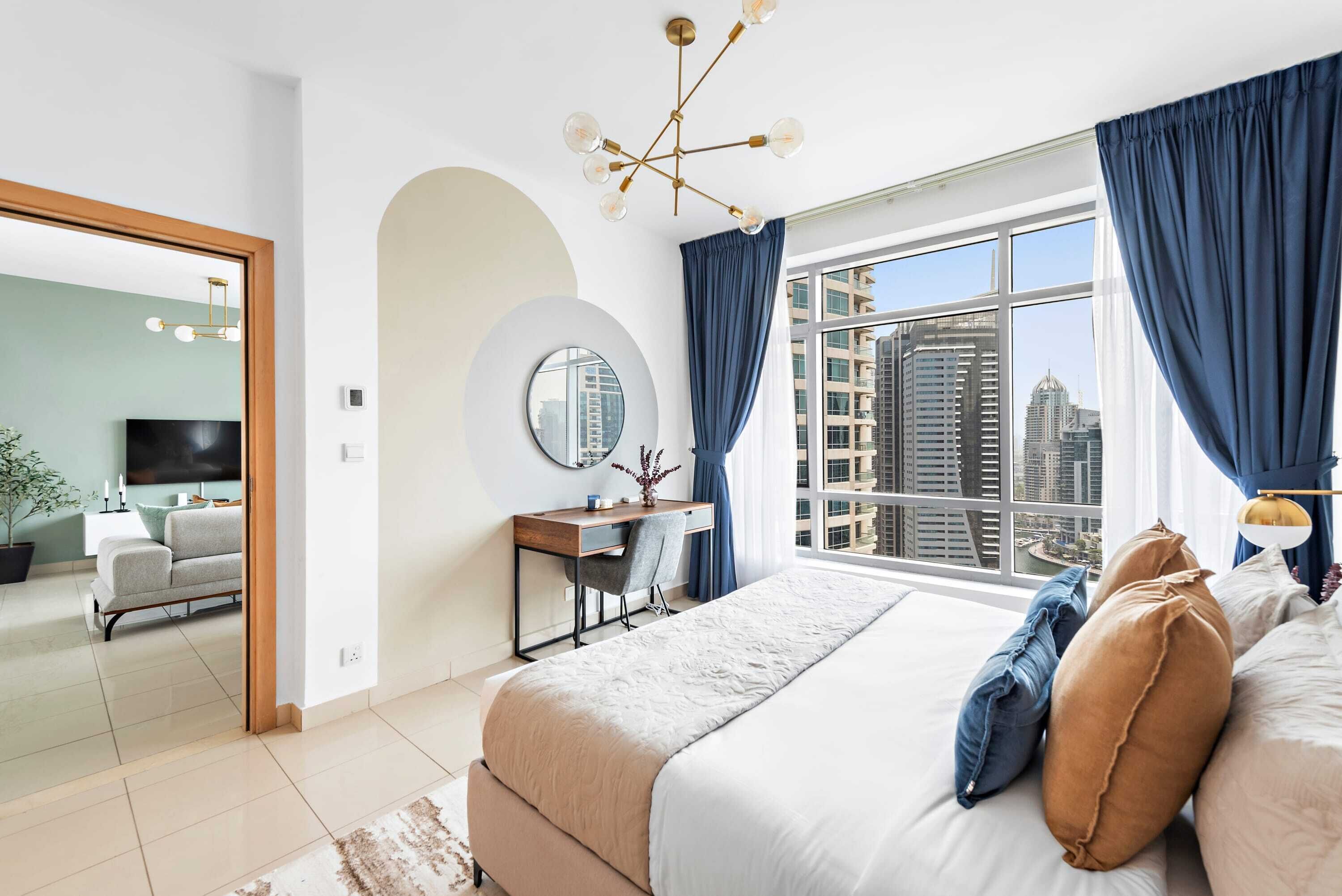 A modern bedroom with a large bed, a desk, a chair, a round mirror, and a view of the city through a large window.