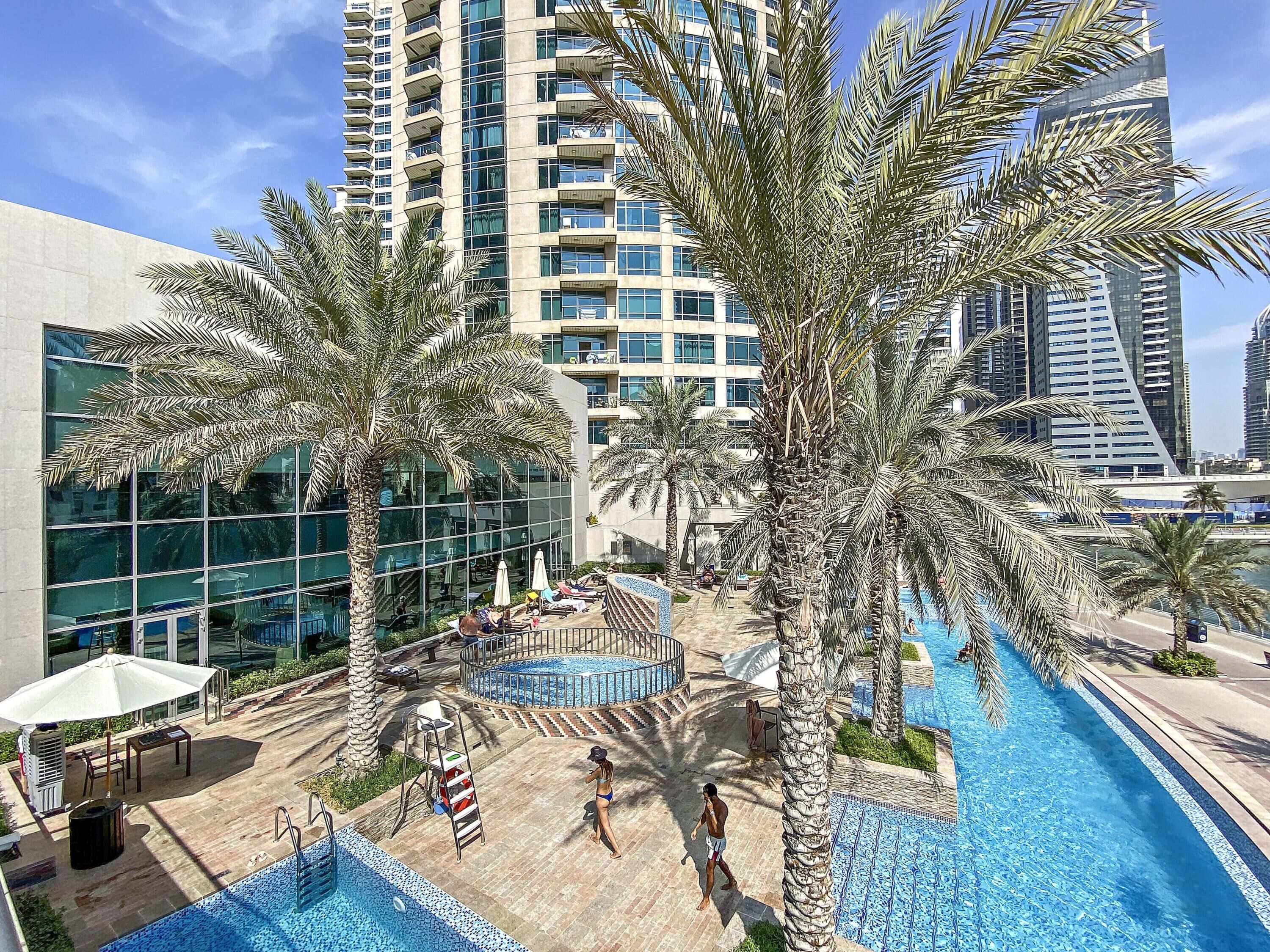 A modern hotel with a rooftop pool, palm trees, and high-rise buildings in the background.