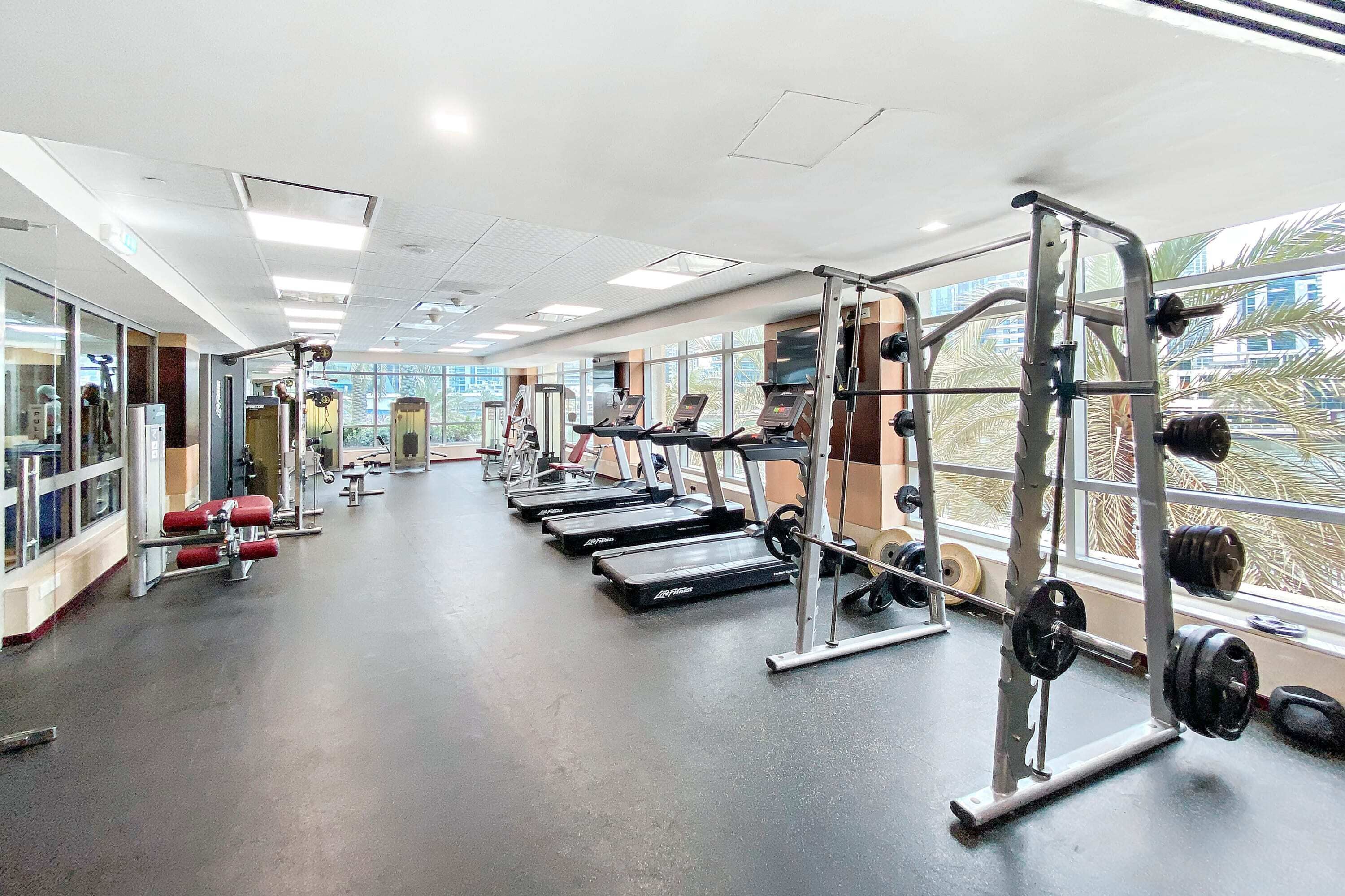 Een sportschool met loopbanden, krachttoestellen en een uitzicht op palmbomen door grote ramen.