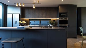 Une cuisine moderne avec des meubles de couleur bois foncé, un grand îlot central doté de tabourets, un saladier rempli de fruits et un luminaire suspendu.