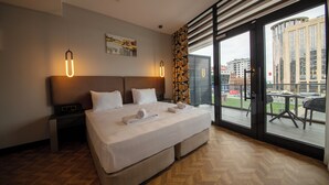 A hotel room with a large bed, a nightstand, a painting on the wall, and a view of the city through large windows. - V Center Hotel (Yomra)