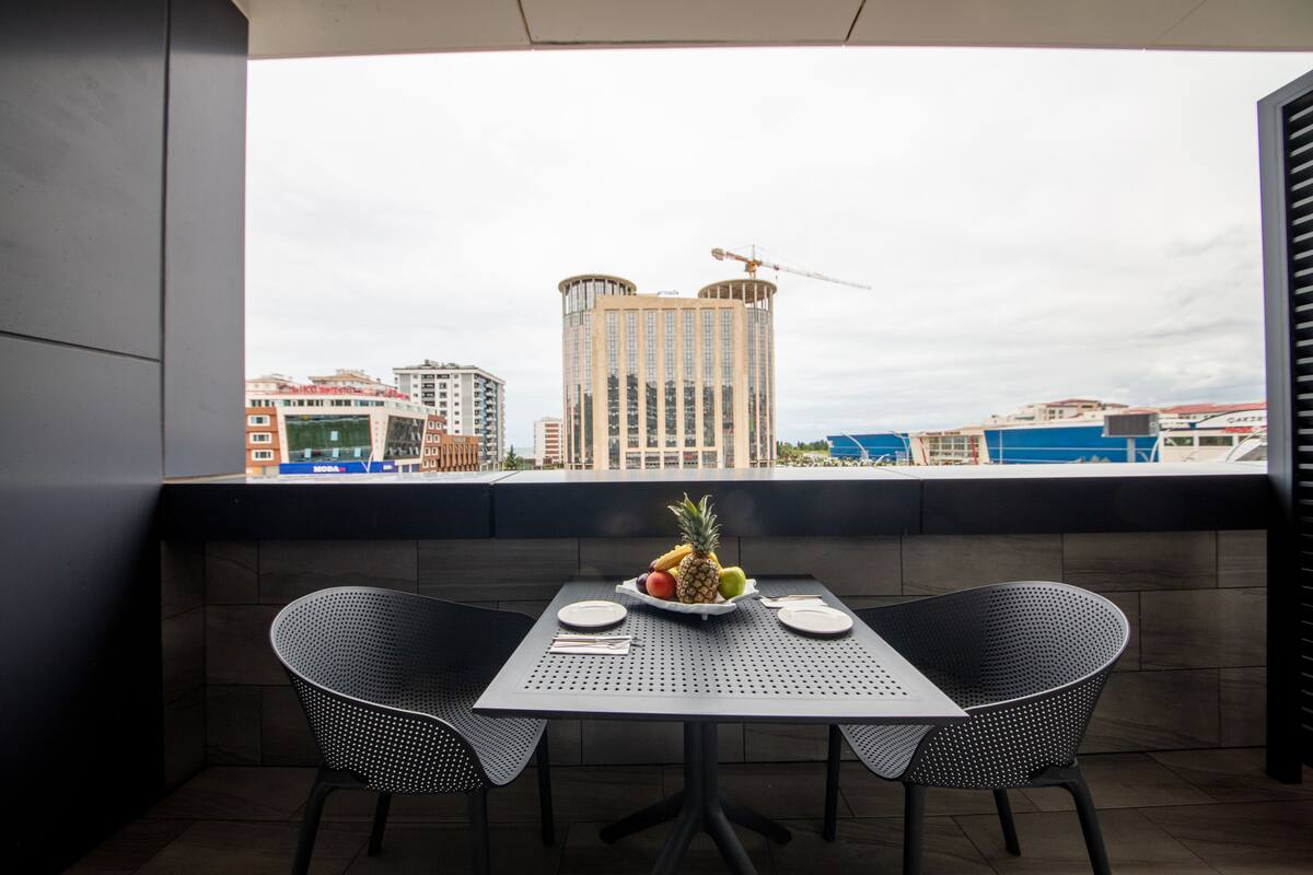 A balcony with two black chairs and a table, a fruit bowl on the table, and a view of buildings and a crane in the background.
